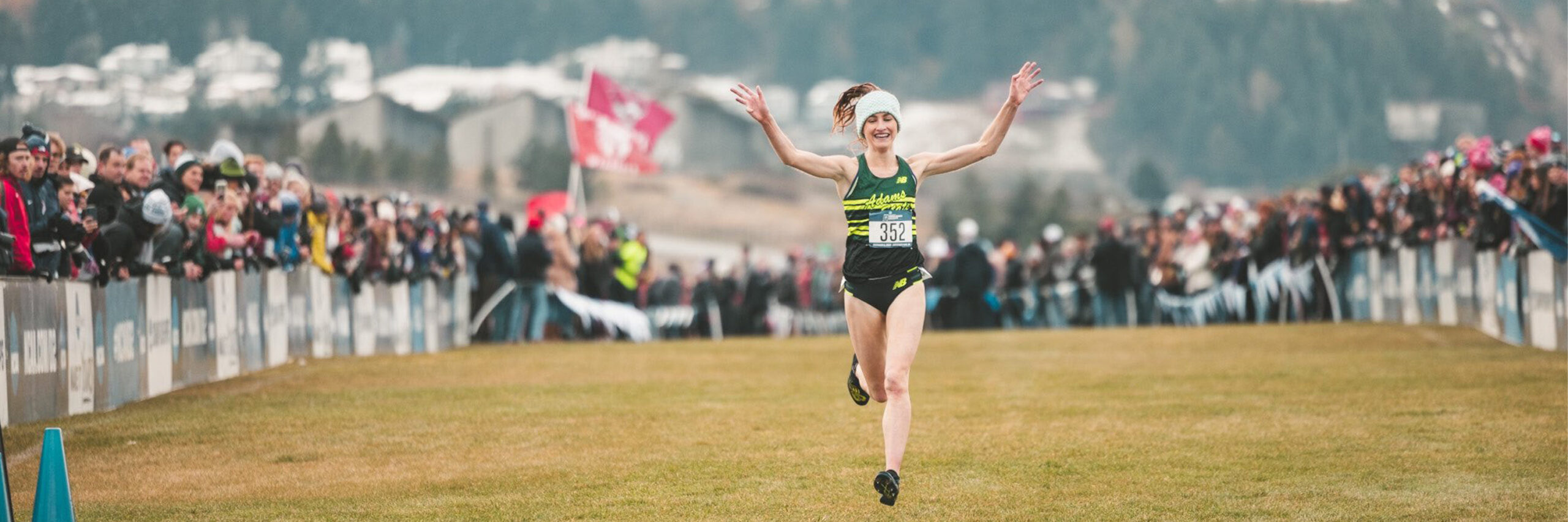 young woman crosses the finish line at a cross-country meet, with spectators on each side.