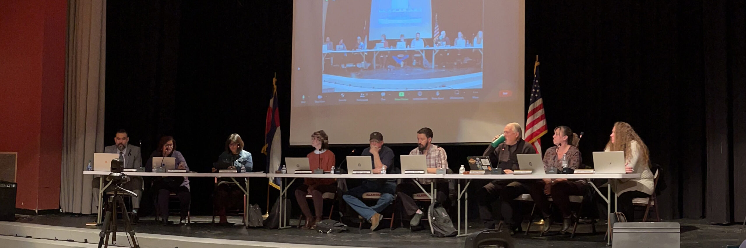 School Board members sitting at a long table on a stage with a screen behind them.